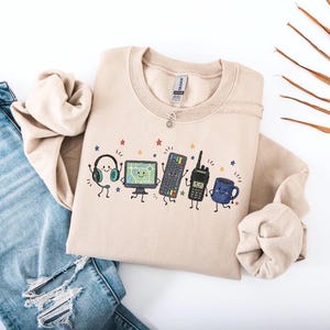 May include: A beige crewneck sweatshirt featuring a graphic print of five anthropomorphic objects: a headset, a computer monitor displaying a map, a keyboard, a walkie-talkie, and a blue coffee mug, all surrounded by small colorful stars.