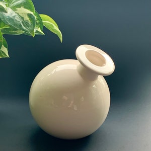 May include: A white ceramic vase with a round body and a wide, open top. The vase is designed with a simple, modern aesthetic.