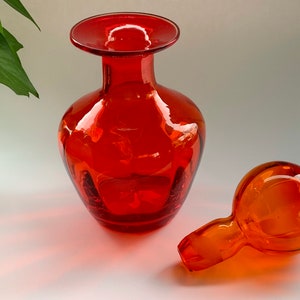 May include: A set of two orange glass decanters with a wide mouth and a round stopper. The decanters are made of clear glass with a slight amber tint.
