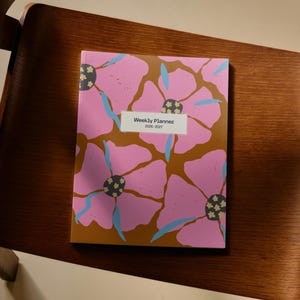 May include: A weekly planner for 2026-2027 featuring a bold floral pattern with large pink petals, dark brown background, and small blue leaves. The center of the cover displays a white rectangular label with the text Weekly Planner 2026-2027.