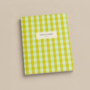 May include: A rectangular weekly planner for the year 2027 featuring a vibrant yellow and light green gingham check pattern. A centered white rectangular label on the front cover displays the black text WEEKLY PLANNER 2027 in a clean font.