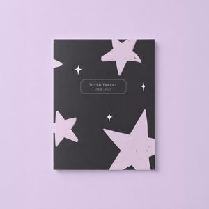 May include: A black weekly planner for 2026-2027 featuring a pattern of light pink hand-drawn stars and small white four-pointed star icons. The center of the cover displays a rectangular label with the text Weekly Planner 2026-2027.