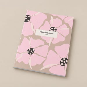 May include: A weekly planner for 2027 featuring a soft pink and beige floral pattern on the cover. The design includes large, stylized light pink flower petals with dark centers containing small white floral accents. A white rectangular label is centered on the front with the black text WEEKLY PLANNER 2027.