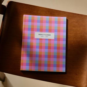 May include: A weekly planner for 2026-2027 featuring a vibrant, multi-colored plaid pattern in shades of pink, orange, and purple. The cover displays a white rectangular label in the center with the black text WEEKLY PLANNER 2026-2027 printed on it.
