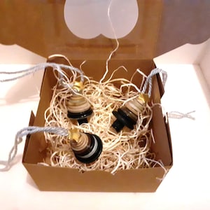 May include: Three decorative bottle stoppers with a layered design, presented in a brown box filled with straw. Each stopper has a black base, a light-coloured body, and a gold star on top. The stoppers are connected by a silver string.