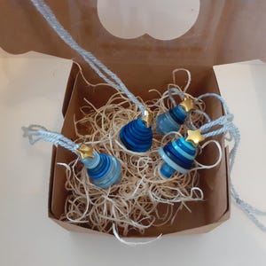 May include: Four blue and white paper ornaments with gold star toppers and silver string, presented in a brown box with straw. The ornaments feature diverse designs, including stripes and solid colours.
