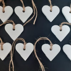 May include: Ten white heart-shaped decorations with a textured finish, each suspended by natural twine. The hearts are arranged on a dark background, creating a rustic, handcrafted look. Ideal for crafting or home decor.