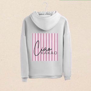 Könnte beinhalten: Weißer Kapuzenpullover mit langen Ärmeln. Auf der Rückseite befindet sich ein rosa-weiß gestreifter Quadrat mit den Worten "Ciao Kakao" in schwarzer Schrift. Der Pullover hängt an einem weißen Kleiderbügel.
