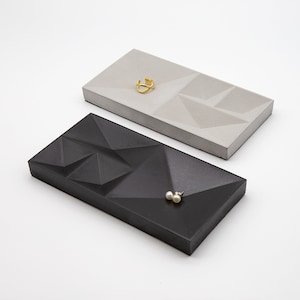 May include: Two geometric concrete trays, one black and one gray, with a gold ring and pearl earrings on top.