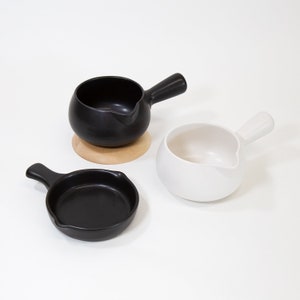 May include: Two small ceramic pitchers with handles, one black and one white, on a white surface. The black pitcher is on a wooden coaster.
