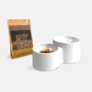 May include: White ceramic double dog bowl with a raised rim. One bowl contains a small amount of brown flakes. The bowl is on a white surface. The text on the brown bag reads "Bonito Flakes Forever".
