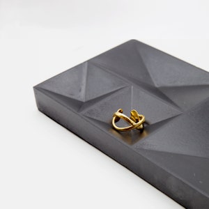 May include: A gold ring with a unique, abstract design sits on a gray concrete tray with a geometric pattern.