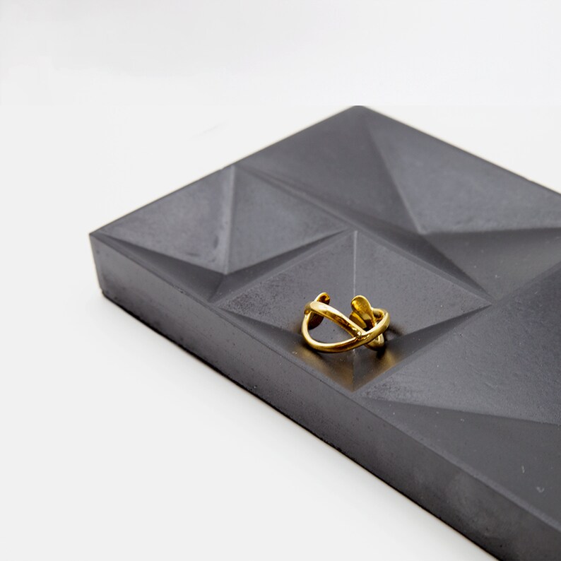 May include: A gold ring with a unique, abstract design sits on a gray concrete tray with a geometric pattern.