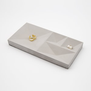 May include: A gray concrete tray with a geometric design. The tray has three compartments, one large and two smaller. A gold ring and a pair of pearl earrings are placed in the compartments.