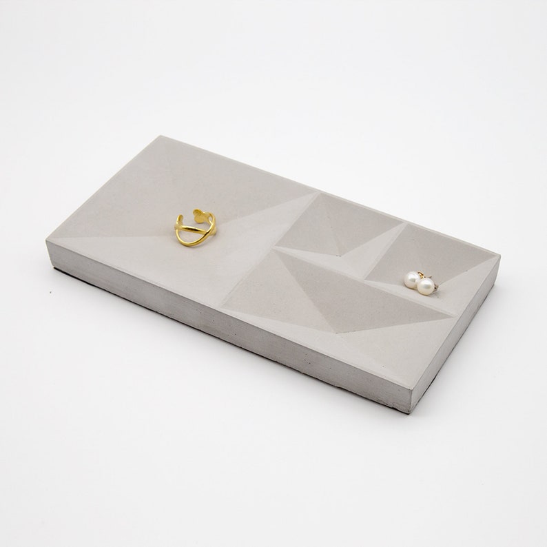 May include: A gray concrete tray with a geometric design. The tray has three compartments, one large and two smaller. A gold ring and a pair of pearl earrings are placed in the compartments.