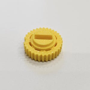 May include: A yellow, gear-shaped object with a rectangular slot in the center. The object has a ridged outer edge and a circular design around the slot. The object is likely a component or part of a larger mechanism.