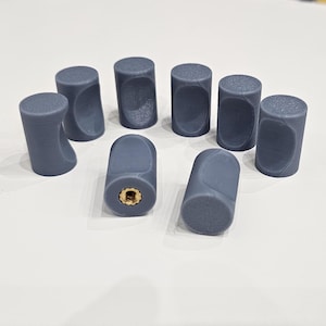 Peut inclure: Ensemble de huit boutons de meuble cylindriques gris. Les boutons ont un dessus arrondi et une poignée incurvée et encastrée sur un côté. Un bouton a un insert fileté de couleur laiton. Les boutons sont disposés sur une surface blanche.