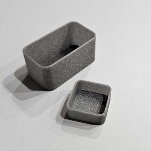 Könnte beinhalten: Zwei rechteckige Behälter mit einer texturierten, metallisch silbernen Oberfläche. Der größere Behälter ist länglich mit abgerundeten Ecken, während der kleinere Behälter quadratisch mit abgerundeten Ecken ist. Beide haben offene Oberseiten.