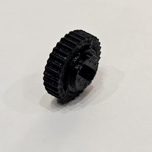 Pode incluir: Uma engrenagem preta impressa em 3D com uma borda externa estriada e um recesso central em forma de cruz. A engrenagem é feita de um material preto fosco e parece ser um componente de um mecanismo maior. A imagem é tirada em um fundo branco liso.
