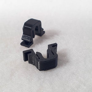 Könnte beinhalten: Zwei schwarze Plastikclips, die jeweils U-förmig sind und an einer Seite eine kleine Lippe haben. Die Clips sind so konzipiert, dass sie Gegenstände an Ort und Stelle halten.