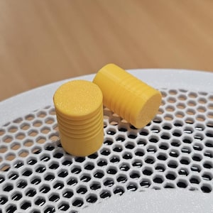 Op de afbeelding: Twee kleine, cilindervormige gele objecten met een getextureerd, plat bovenoppervlak en horizontale gegroefde lijnen langs de zijkanten. Deze items zijn geplaatst op een wit, rigide oppervlak met een herhalend zeshoekig rasterpatroon.