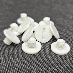Op de afbeelding: Een verzameling witte plastic schroeven met een platte, ronde kop en een schroefdraad. De schroeven liggen verspreid op een grijze, getextureerde ondergrond, waarbij sommige scherp en andere enigszins wazig zijn.