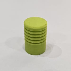 Może przedstawiać: Cylindryczny, limonkowo-zielony przedmiot z poziomymi rowkami. Wierzch to płaskie, pełne koło. Obiekt jest w jednolitym kolorze i wydaje się być wykonany z tworzywa sztucznego lub podobnego materiału. Obiekt leży na białej powierzchni.