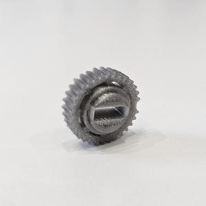 Può includere: Un piccolo ingranaggio grigio stampato in 3D con un bordo esterno dentato e un foro quadrato al centro. L'ingranaggio ha una superficie strutturata e dettagliata ed è probabilmente realizzato in plastica. L'oggetto è posto su uno sfondo bianco.