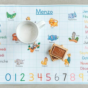 Peut inclure: Un set de table blanc à motif quadrillé, avec des illustrations colorées d'animaux et de chiffres. Le set de table porte le nom "Menzo" en haut, avec une tasse de lait et deux biscuits dessus.