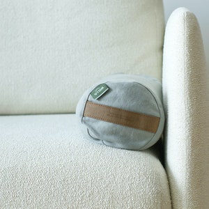 May include: A light gray cylindrical pillow with a brown leather strap and a green label that says "WILO" on a white couch.