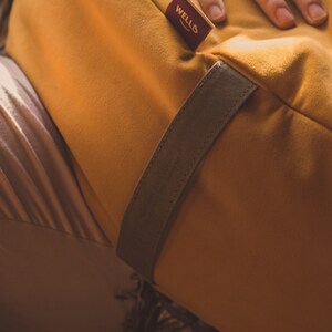 A mustard yellow crescent-shaped meditation cushion with a soft suede-like fabric, featuring a brown carrying strap and a WELLB brand tag. A relaxed hand rests on the cushion, creating a cozy, warm ambiance. Perfect for yoga, mindfulness.