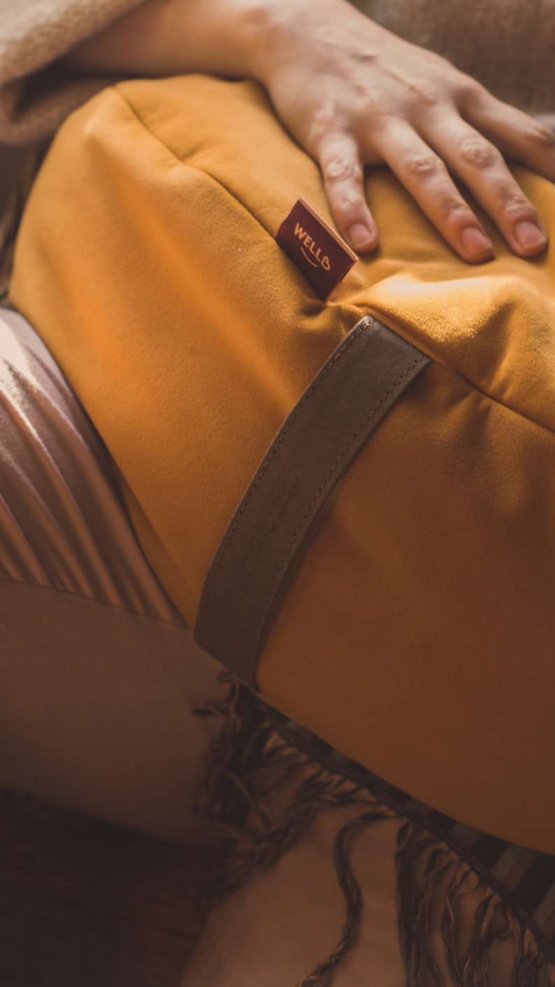 A mustard yellow crescent-shaped meditation cushion with a soft suede-like fabric, featuring a brown carrying strap and a WELLB brand tag. A relaxed hand rests on the cushion, creating a cozy, warm ambiance. Perfect for yoga, mindfulness.