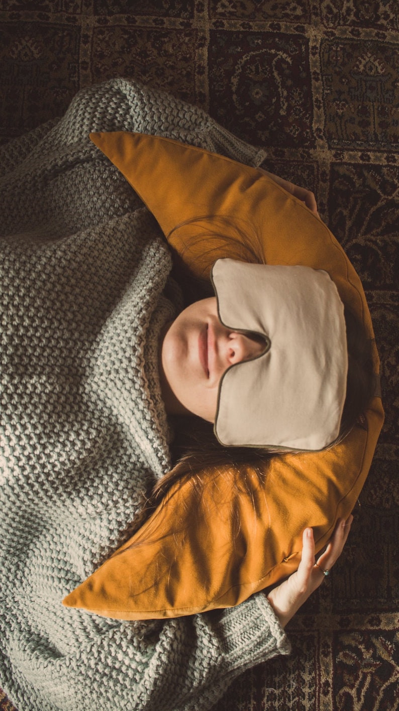 A woman relaxes on a patterned rug, wrapped in a gray blanket. She rests her head on a mustard yellow crescent-shaped pillow while wearing a soft beige eye pillow with a nose cutout. A cozy and calming setup for meditation, rest, and relaxation.