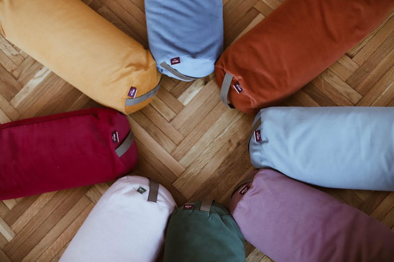 May include: Eight cylindrical yoga bolsters in various colors, including red, blue, beige, white, green, and orange. The bolsters are arranged in a circle on a wooden floor.