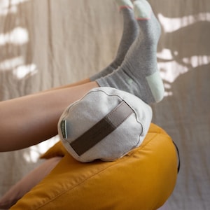 A person rests their legs on a stack of WELLB meditation cushions, wearing cozy gray socks. A small, round linen bolster with a brown carrying strap and a "WELLB" tag supports their ankles. The cushions in mustard yellow create a warm, relaxing setup