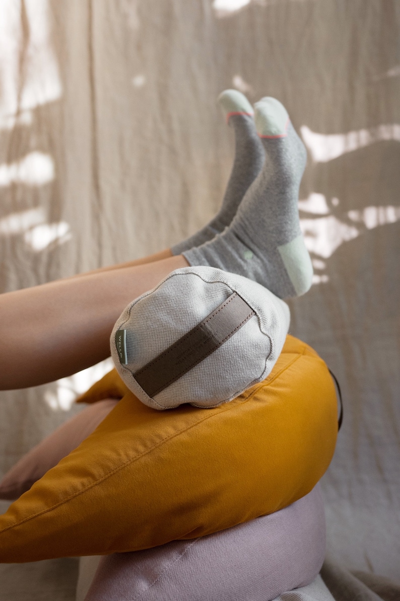 A person rests their legs on a stack of WELLB meditation cushions, wearing cozy gray socks. A small, round linen bolster with a brown carrying strap and a "WELLB" tag supports their ankles. The cushions in mustard yellow create a warm, relaxing setup