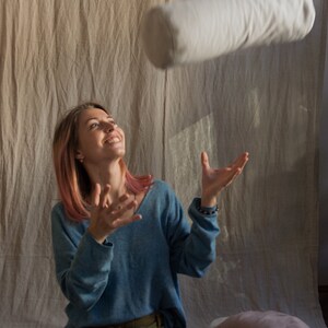 May include: A woman with pink hair is catching a white cylindrical pillow in mid-air. She is wearing a blue sweater and a green skirt. There are two pillows on the floor behind her.