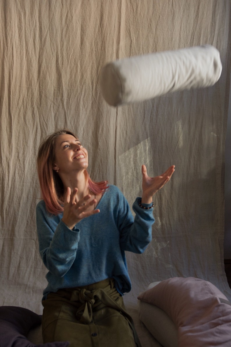 May include: A woman with pink hair is catching a white cylindrical pillow in mid-air. She is wearing a blue sweater and a green skirt. There are two pillows on the floor behind her.