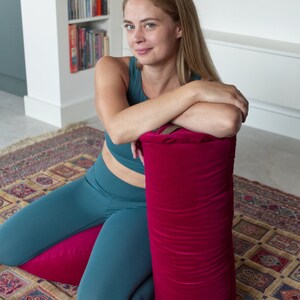 May include: A woman sits on a patterned rug with her legs crossed. She is wearing teal leggings and a teal sports bra. She is leaning against a large, bright red cylindrical pillow.