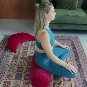 May include: A woman sits cross-legged on a red meditation cushion, with a red crescent-shaped cushion behind her. She is wearing a teal tank top and teal leggings. The cushions are on a patterned rug.