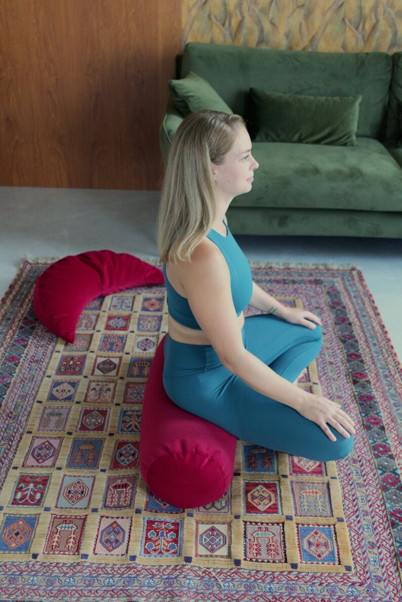 May include: A woman sits cross-legged on a red meditation cushion, with a red crescent-shaped cushion behind her. She is wearing a teal tank top and teal leggings. The cushions are on a patterned rug.