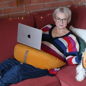 May include: A woman wearing glasses and a striped sweater sits on a red couch with a laptop. An orange pillow supports the laptop. A star-shaped decoration with lights hangs on the brick wall behind her. A white and blue floral teacup sits on a wooden table.