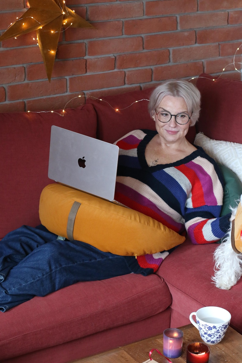 May include: A woman wearing glasses and a striped sweater sits on a red couch with a laptop. An orange pillow supports the laptop. A star-shaped decoration with lights hangs on the brick wall behind her. A white and blue floral teacup sits on a wooden table.