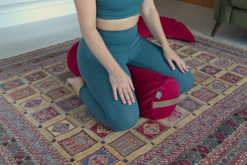 May include: A person sits on a red meditation cushion with a brown strap. The cushion is on a patterned rug.