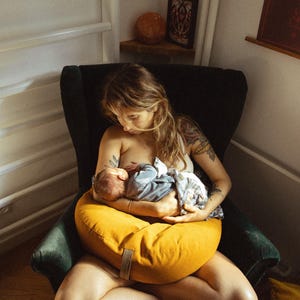 May include: A woman sits in a green armchair, breastfeeding a baby while using a yellow nursing pillow. The woman has long brown hair and is wearing a white tank top. The baby is wrapped in a blue blanket.