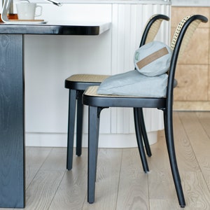 May include: Two black chairs with woven cane seats and backs. The chairs have gray cushions and a brown pillow on the seat of one chair. The chairs are in front of a white kitchen counter and a wooden cabinet.