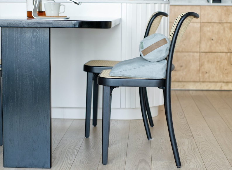 May include: Two black chairs with woven cane seats and backs. The chairs have gray cushions and a brown pillow on the seat of one chair. The chairs are in front of a white kitchen counter and a wooden cabinet.