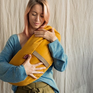 A woman with soft pink hair and a gentle smile embraces a mustard yellow WELLB crescent-shaped meditation cushion. She wears a cozy blue sweater and an olive green tied-waist skirt. A warm and calming wellness accessory for relaxation.
