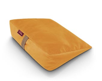 May include: Mustard-colored wedge pillow with a soft, velvety texture. The pillow has a rectangular shape with a slanted top and a brown strap. The word "WELLO" is printed on a small tag.