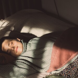 May include: A person sleeping in a bed with a white blanket and a pink pillow. The person is wearing a light green sweater and has dark hair.